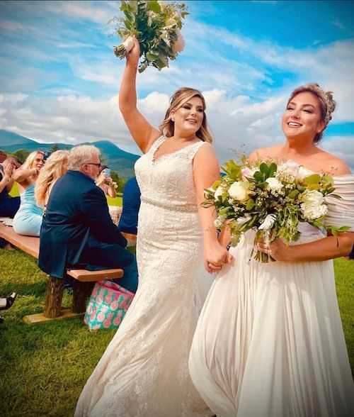 Two brides celebrate outdoors with bouquets, guests seated on benches, mountains in background.