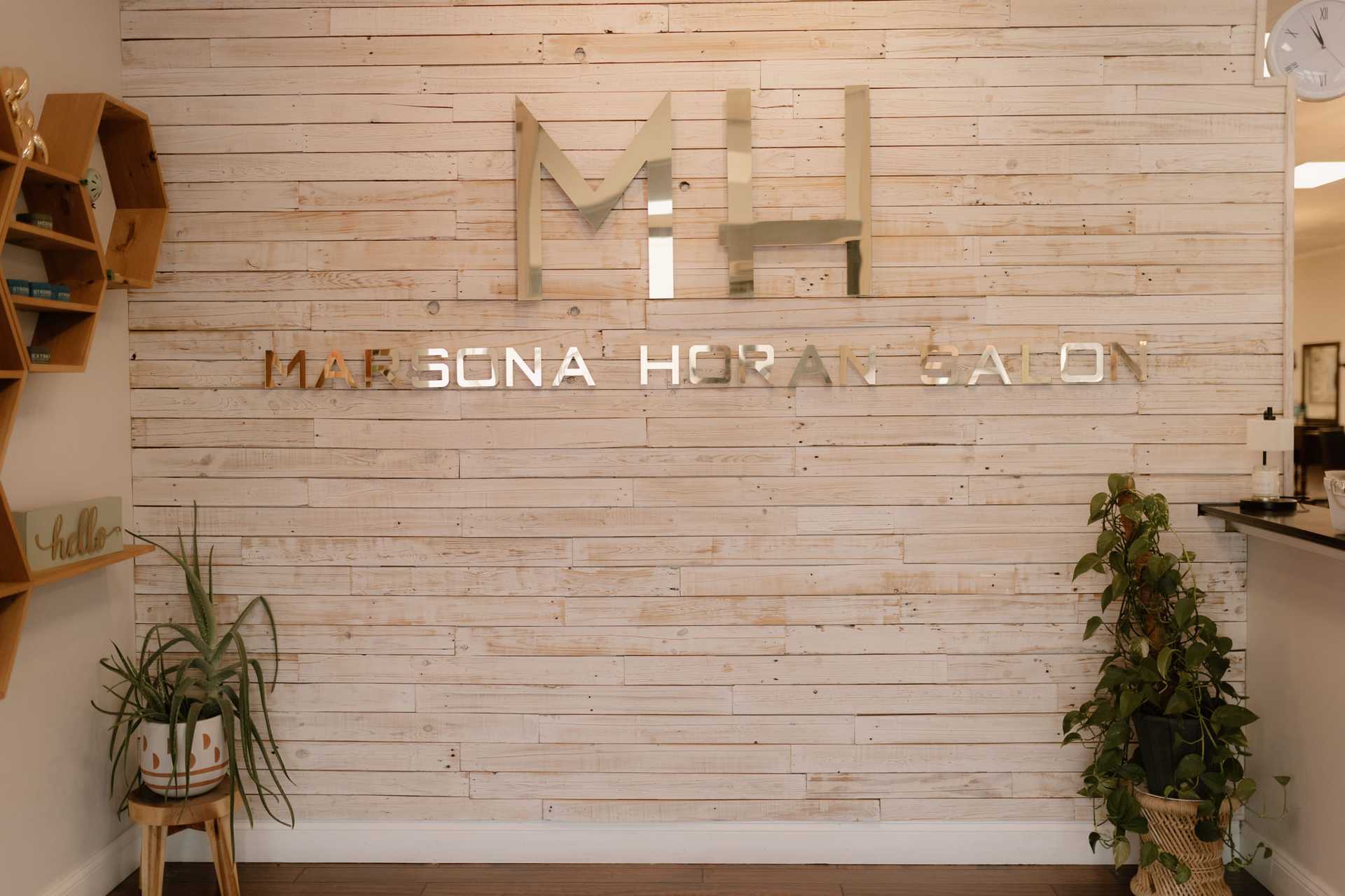 ```plaintext
Mae Soná Hair Salon sign on a wooden wall with plants and shelving.
```